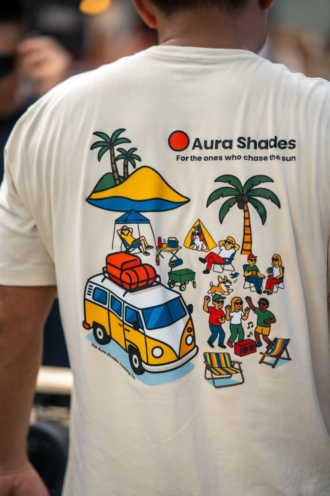 Person wearing t-shirt with colorful summer beach scene, camper van, palm trees, and people relaxing.